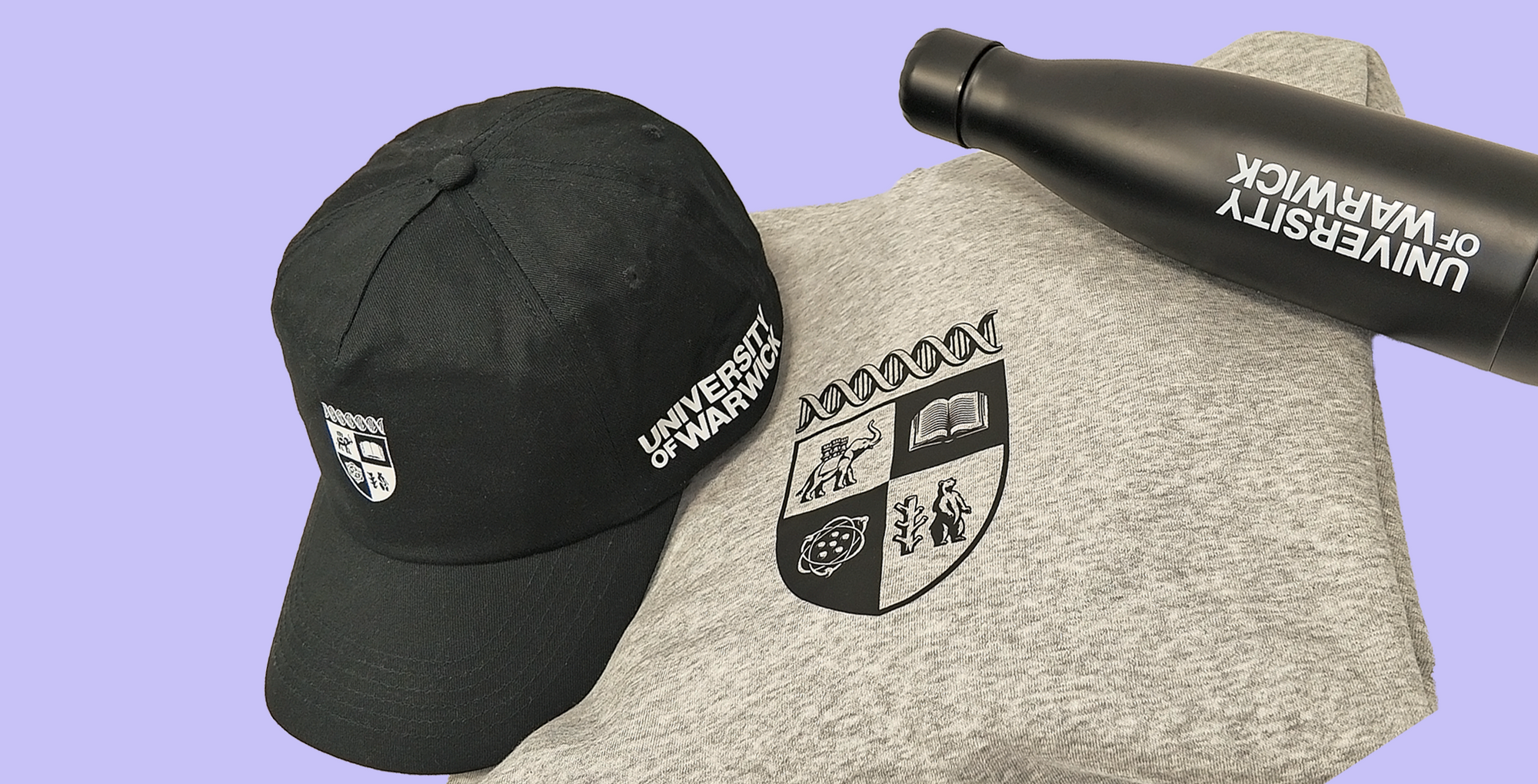 A black baseball cap and a black insulated bottle with white University of Warwick branding, arranged on top of a heather gray T-shirt featuring the university crest, against a solid lavender background.