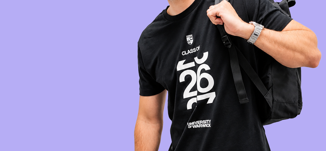 A man wearing a black t-shirt with a white graphic print that reads 'CLASS OF 2026 UNIVERSITY OF WARWICK' and carrying a black backpack against a solid purple background.