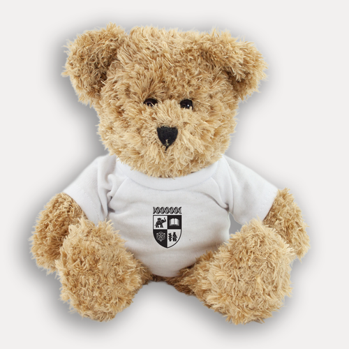 A plush teddy bear with light brown fur wearing a white short-sleeve T-shirt with a black University of Warwick crest graphic on the chest, shown against a light gray background.